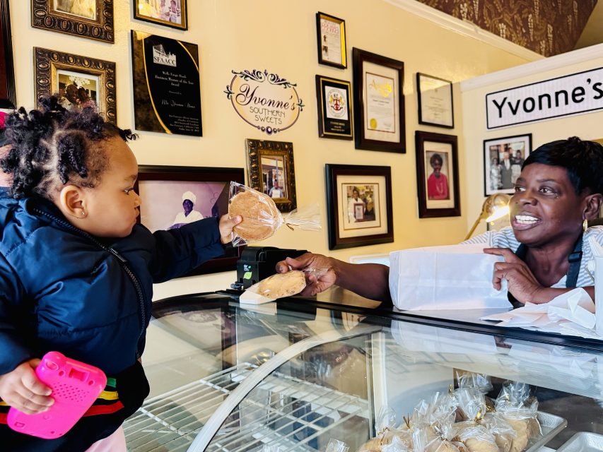 A woman behind a glass display case hands a packaged cookie to a young child holding a pink toy inside a bakery decorated with awards and family photos.