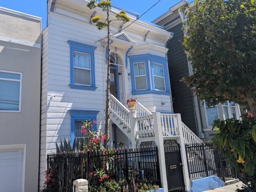 A two-story white house with blue trim, a bay window, and a staircase leading to the front door, surrounded by a black metal fence and plants.