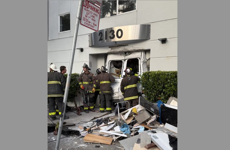 Firefighters stand near a damaged building entrance with debris scattered on the sidewalk after a vehicle crashed into the front of the structure at address 12130.