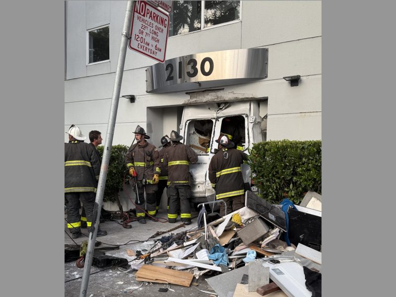 Firefighters stand near a damaged building entrance with debris scattered on the sidewalk after a vehicle crashed into the front of the structure at address 12130.