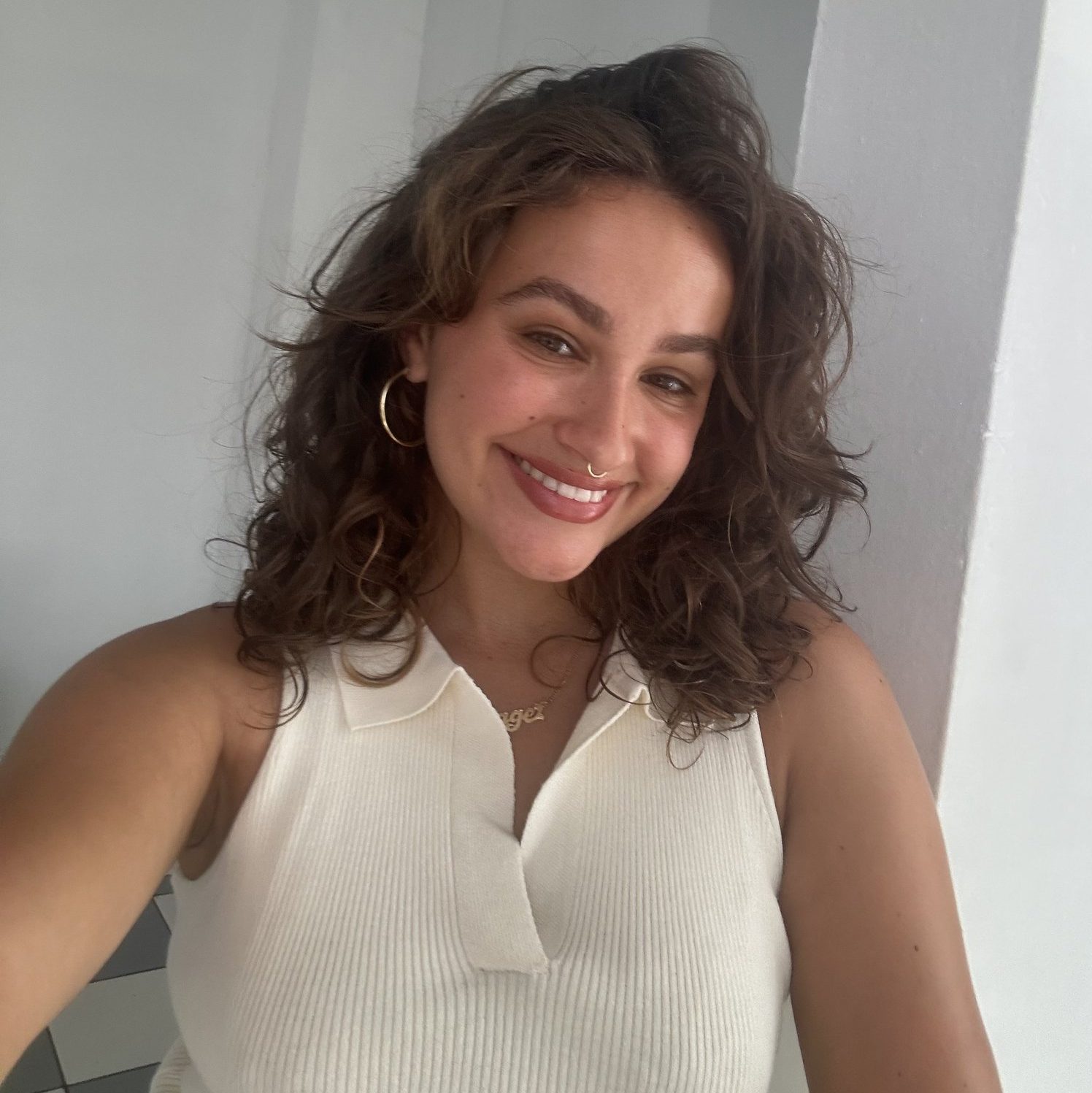 A woman with wavy brown hair, wearing a sleeveless white top, gold hoop earrings, and a necklace, smiles at the camera indoors.