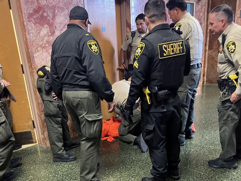 Several uniformed sheriff deputies surround and detain a person in an orange outfit on the floor near a doorway inside a building with marble walls.
