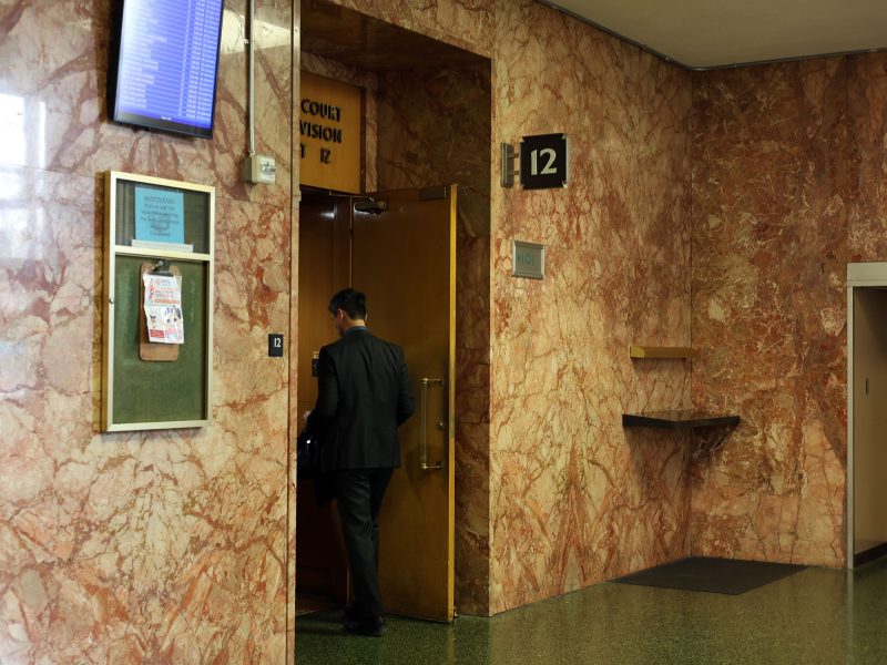 A man in a suit enters a door labeled "Court Division 12" in a marble-walled hallway with a bulletin board and sign displaying the number 12.