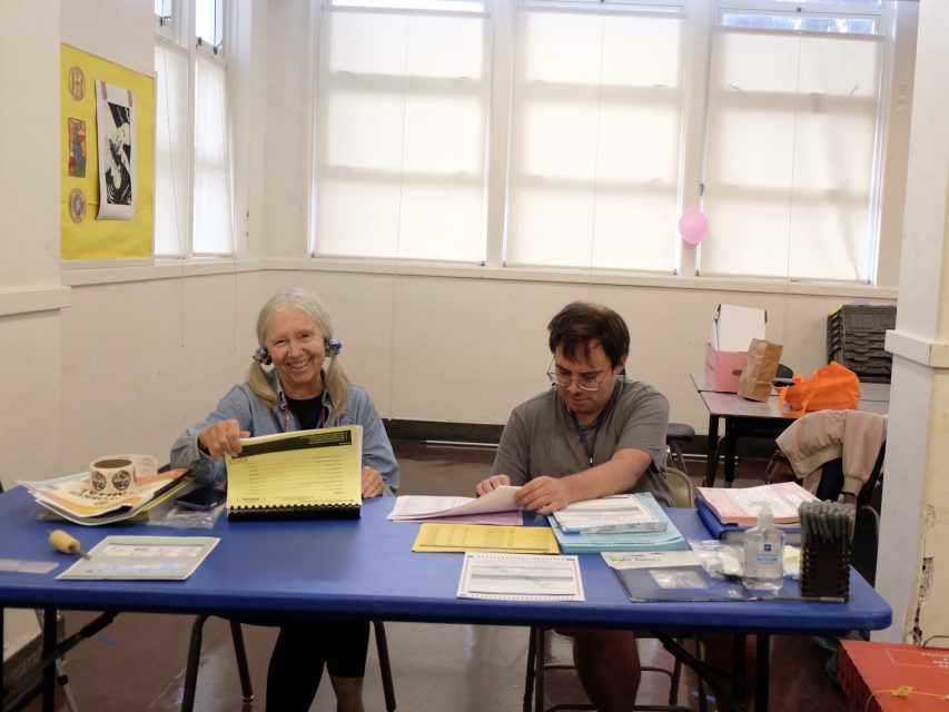 Two people sit at a blue table in a classroom, organizing papers and documents. The woman on the left smiles, while the man on the right focuses on paperwork.