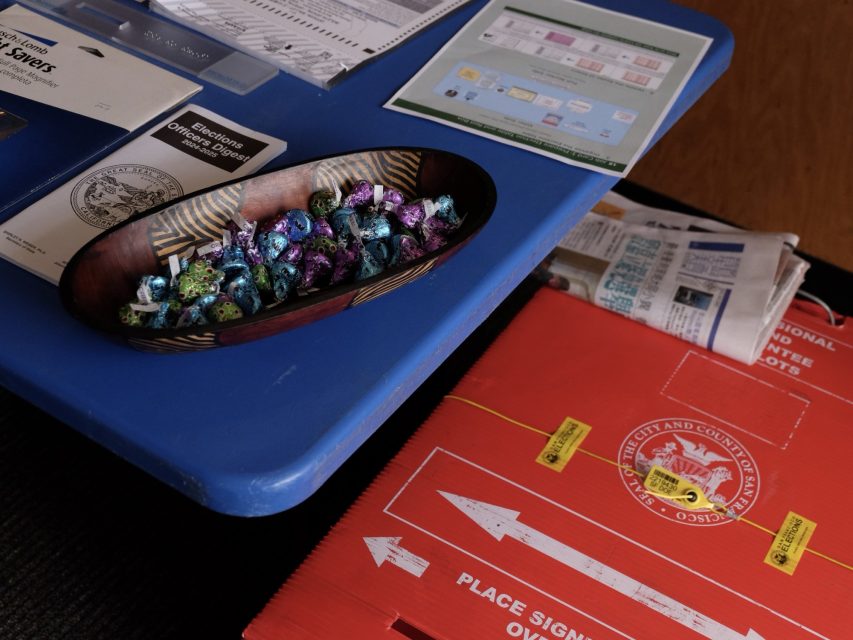 A table with voting materials, a bowl of wrapped candy, and a red ballot drop-off box on the floor, labeled for the City and County of San Francisco.