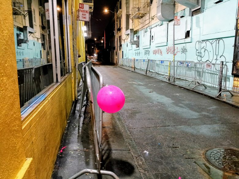 A bright pink balloon is tied to a metal barricade on a graffiti-lined, empty alley at night.