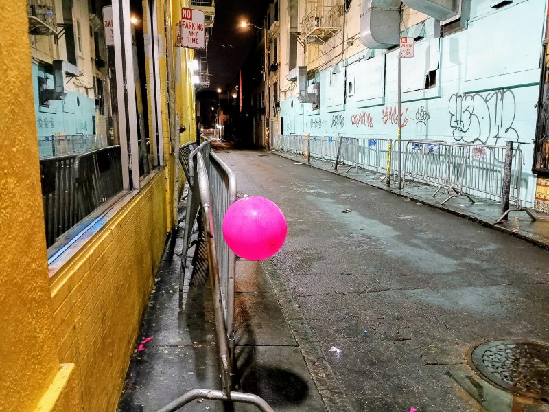 A bright pink balloon is tied to a metal barricade on a graffiti-lined, empty alley at night.