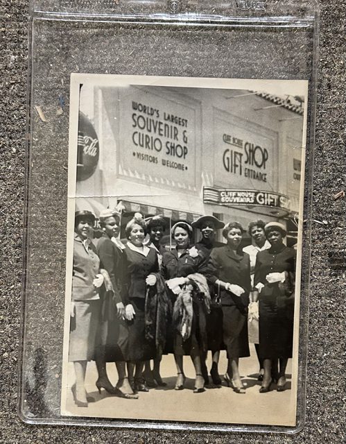 A group of women in formal attire poses in front of a souvenir and curio shop with large signs, on a city sidewalk.