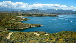 A large lake with blue water surrounded by green hills and a winding dirt road under a partly cloudy sky, with mountains in the background.