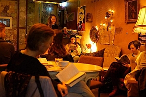 A group of people sit in a cozy, dimly lit room, reading books and talking, with warm lighting and eclectic decorations on the walls.