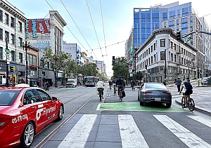 Urban street scene with cars, cyclists in a bike lane, pedestrians crossing, and buildings lining both sides of the road.
