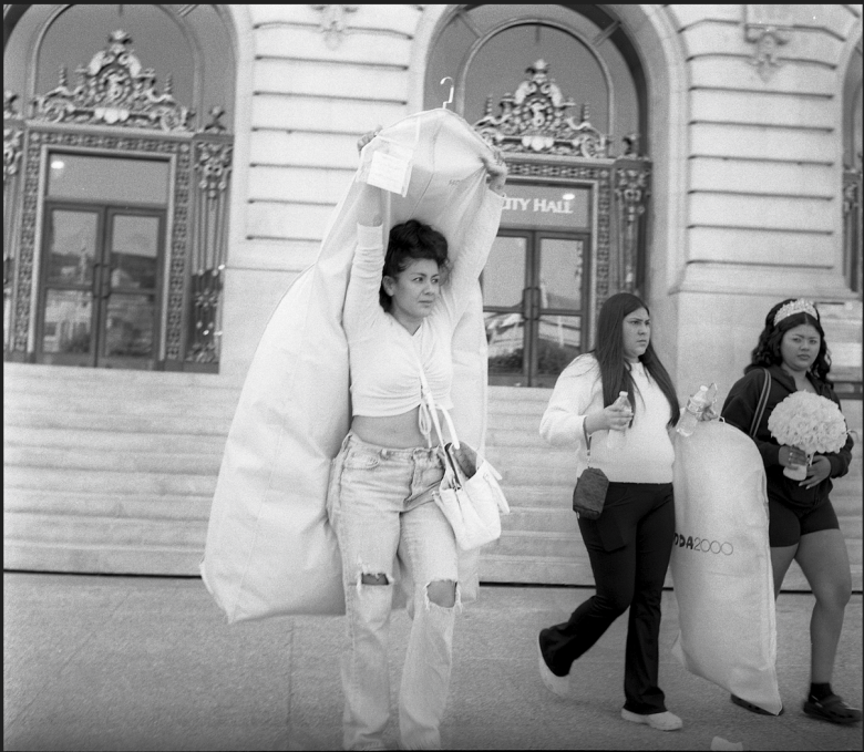 Three women walk in front of a city hall building; one carries a garment bag over her head, another holds a bag, and the third carries a bouquet.
