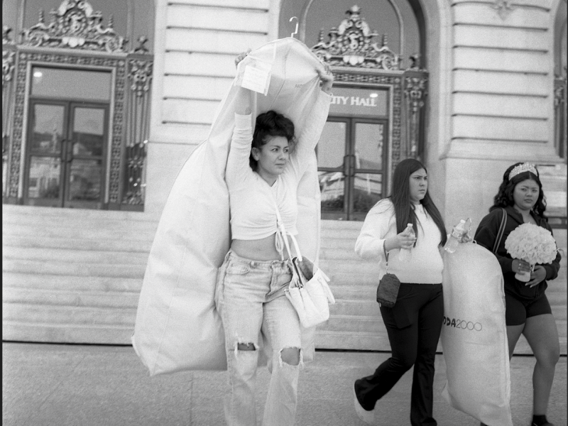 Three women walk in front of a city hall building; one carries a garment bag over her head, another holds a bag, and the third carries a bouquet.