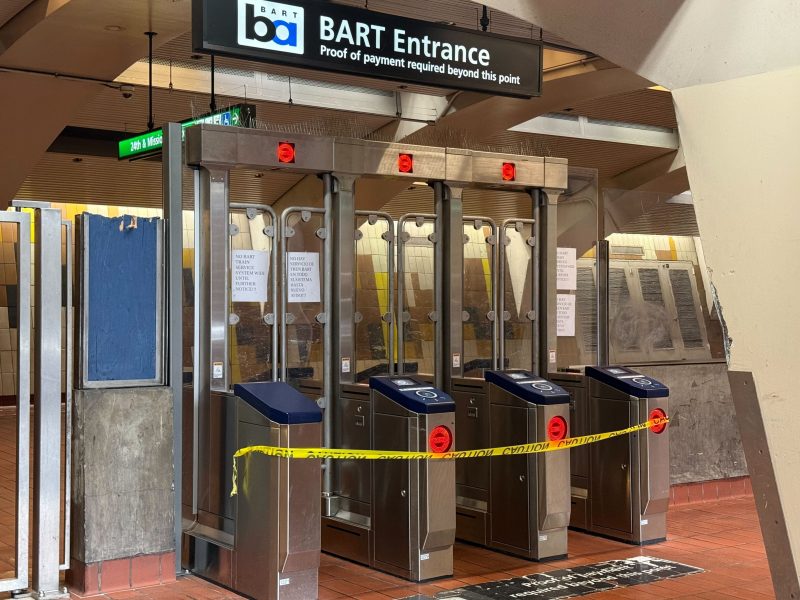 BART station entrance with turnstiles blocked off by caution tape, preventing access. Signs indicate proof of payment is required beyond this point.