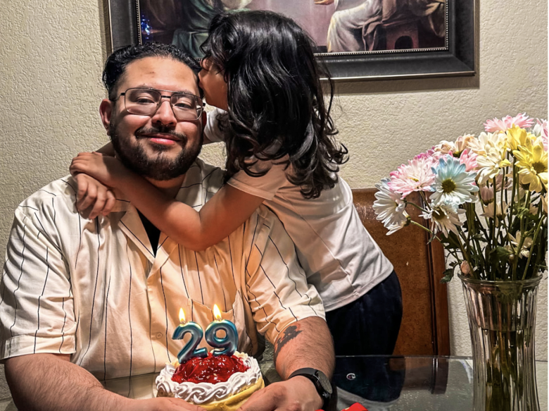 A man sits at a table with a small birthday cake showing the number 29, while a child hugs and whispers to him. A vase of flowers is on the table.