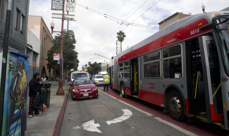 A red SFMTA bus stopped in a designated bus lane next to a red car, with people standing nearby and buildings lining the street.