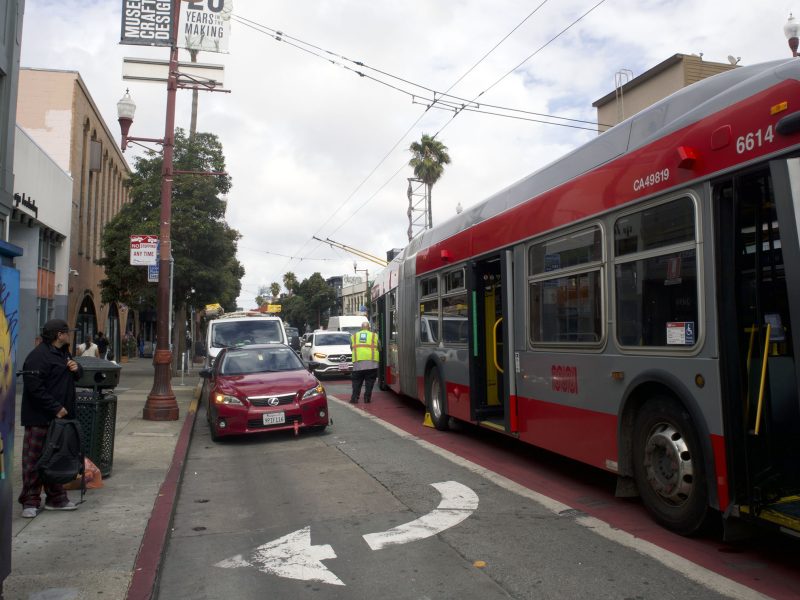 A red SFMTA bus stopped in a designated bus lane next to a red car, with people standing nearby and buildings lining the street.