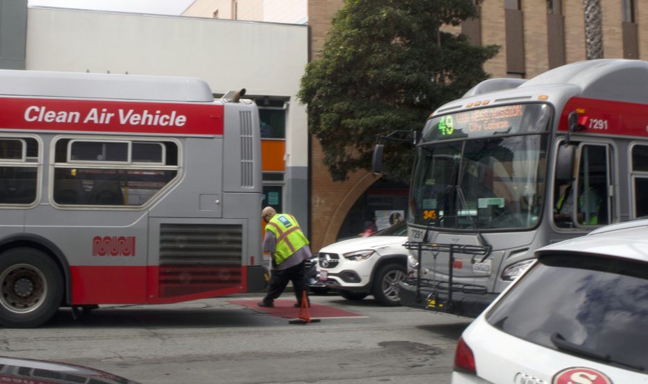 A worker in a reflective vest stands between two city buses on a street as traffic cones block part of the road.