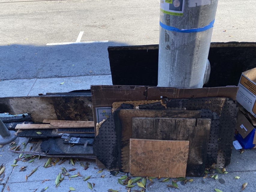 Several pieces of wood and cardboard, some burnt or damaged, are stacked against a utility pole on a sidewalk next to fallen leaves and a curb.