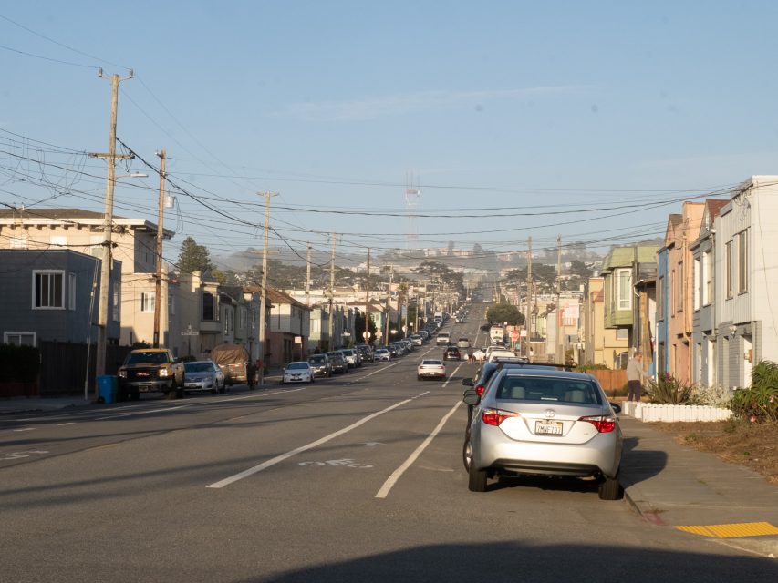 A wide street in a residential neighborhood with parked cars on both sides and houses lining the road; utility poles and wires overhead.