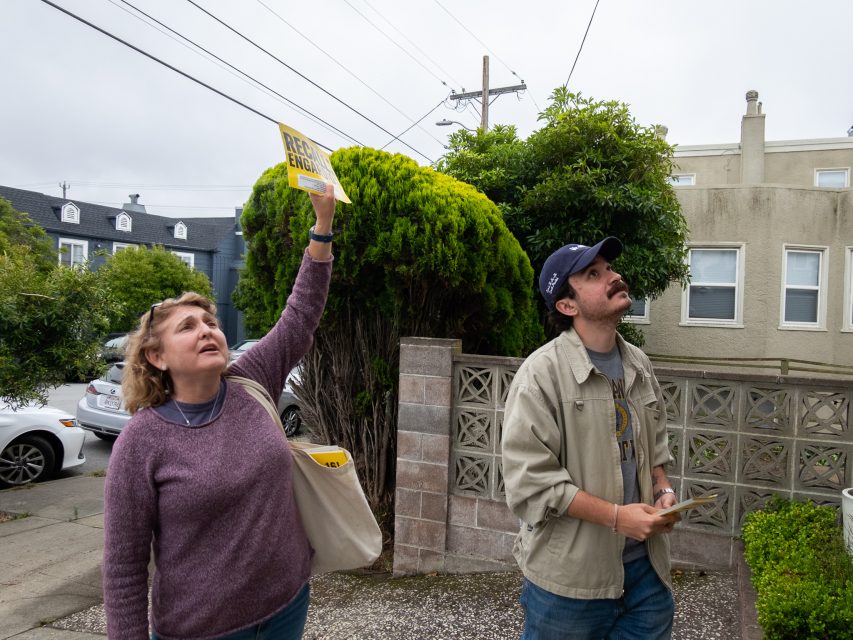 A woman holds up a yellow sign while standing outside with a man. Both are looking up, near a sidewalk and residential buildings.