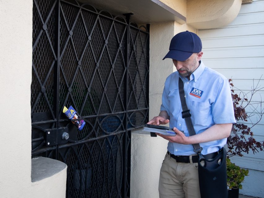 A man in a blue uniform and cap stands outside a gated door, using a tablet. A campaign flyer is clipped to the gate.