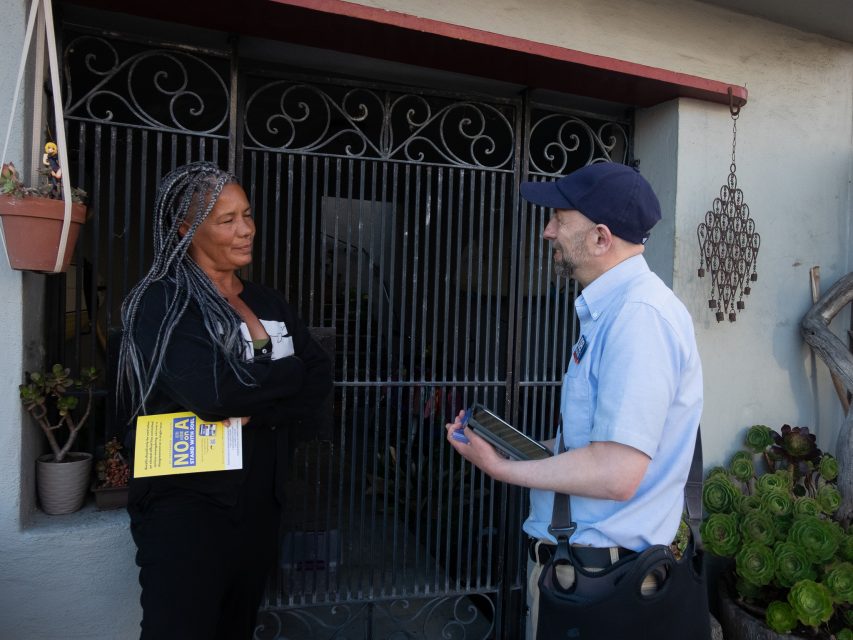 A person with braided hair stands at a doorway holding a flyer while talking to a person in a cap and uniform holding a clipboard.