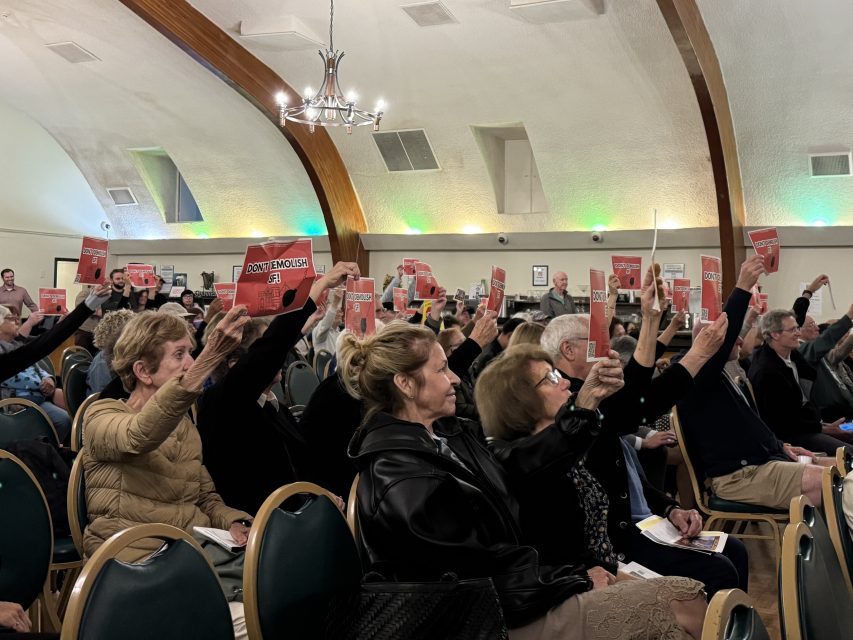 A group of people seated in a hall raise red voting cards during a meeting or assembly.