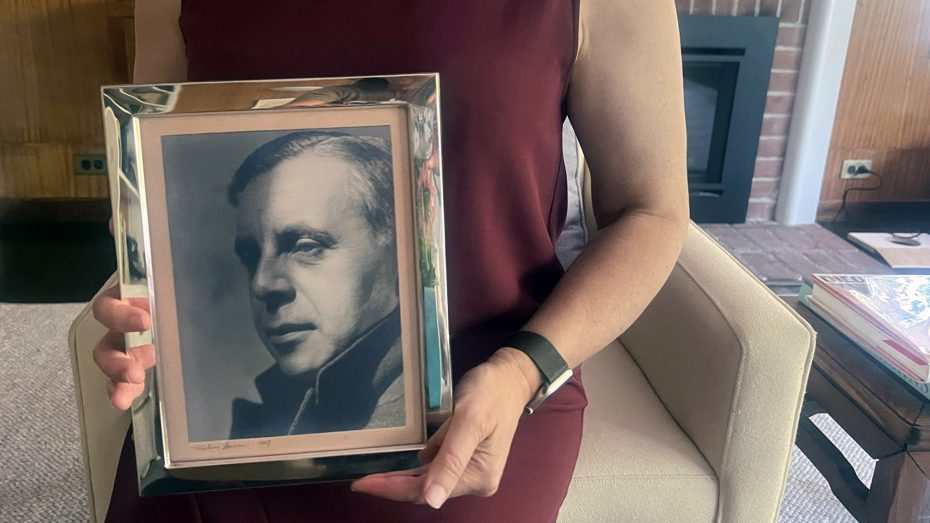 A person in a burgundy dress sits in a chair, holding a framed black-and-white portrait of Diego Rivera.