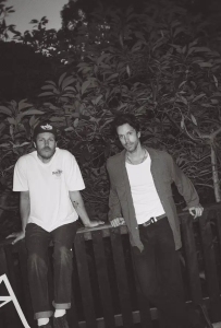 Two men pose outdoors by a wooden fence, one sitting and one standing, with trees and foliage in the background. The image is in black and white.