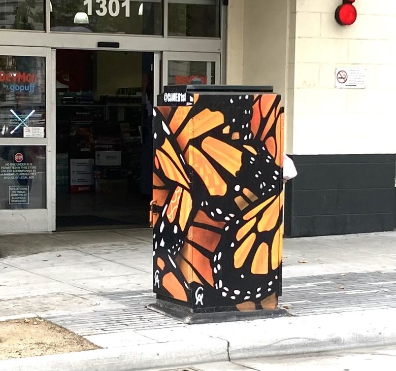 A utility box on a city sidewalk is painted with an orange and black monarch butterfly wing design, positioned near the entrance of a store.