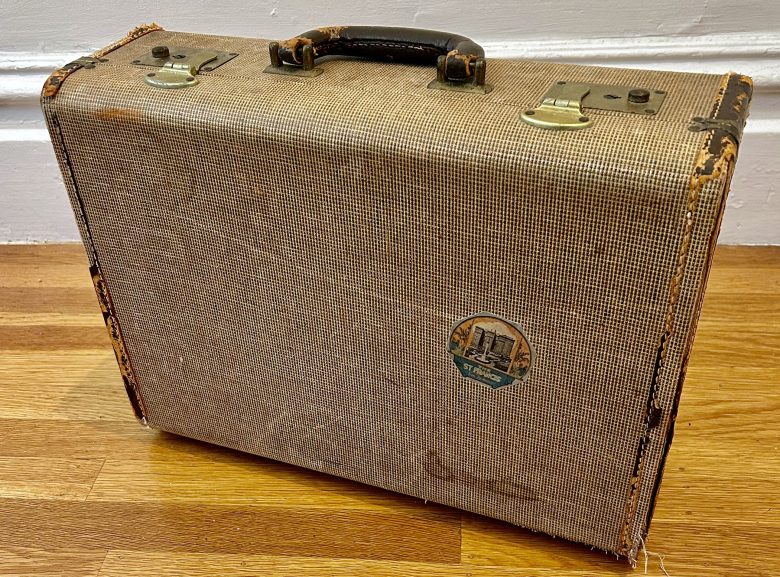 A vintage, tan-colored suitcase with worn edges, metal clasps, a black handle, and a round travel sticker, resting on a wooden floor against a white wall.