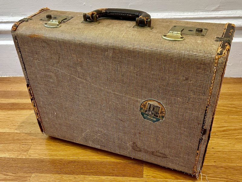 A vintage, tan-colored suitcase with worn edges, metal clasps, a black handle, and a round travel sticker, resting on a wooden floor against a white wall.