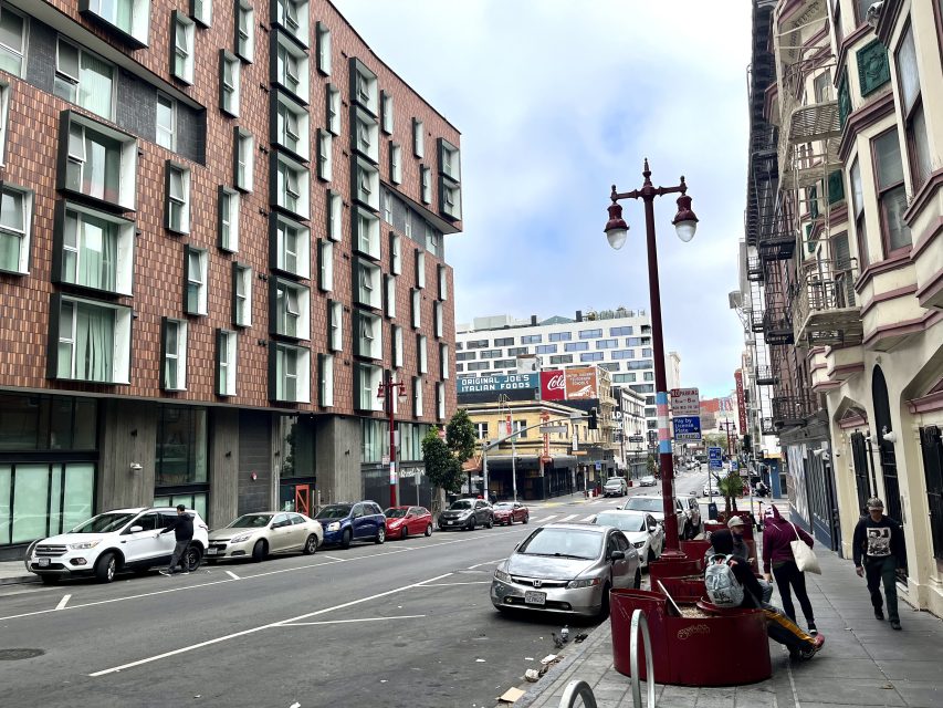 A city street lined with parked cars, modern and older buildings, pedestrians on the sidewalk, and cloudy sky overhead.