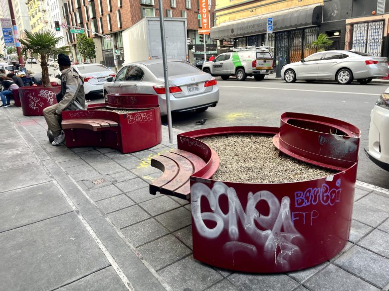 Red circular benches with planters, some covered in graffiti, line a city sidewalk; a few cars are parked on the street and a person sits on one of the benches.