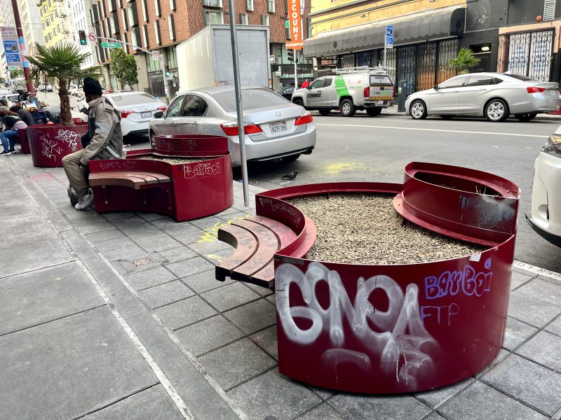 Red circular benches with planters, some covered in graffiti, line a city sidewalk; a few cars are parked on the street and a person sits on one of the benches.