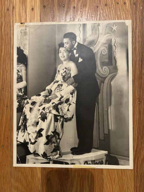 A woman in a floral dress sits on a platform while a man in a tuxedo stands beside her. They pose in front of a decorative backdrop on a wooden floor.