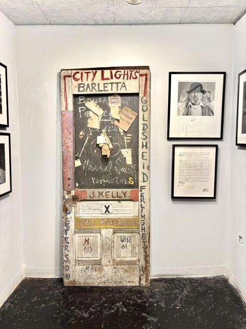 An old, painted door with handwritten names and notes displayed upright in a gallery, surrounded by framed photographs and documents on white walls.
