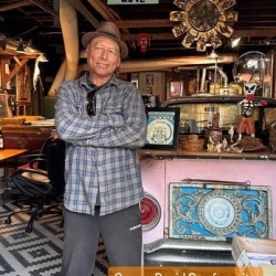 An older man in a hat and plaid shirt stands with crossed arms in an eclectic, art-filled room with various decor and collectibles.