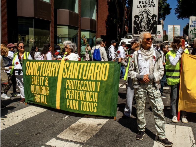 A group of people participate in a street march holding banners reading "Sanctuary Safety & Belongings For All" and "Santuario Protección y Pertenencia Para Todos.