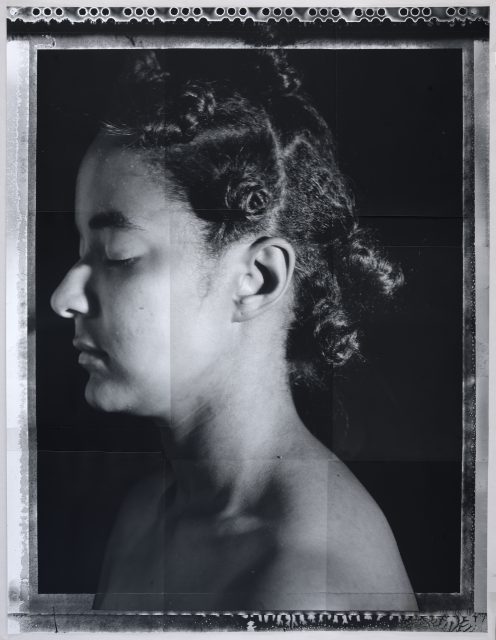 A black and white portrait of a person with closed eyes in profile view, showing short curled hair against a dark background.