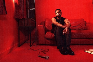 A man sits on a red couch in a red-lit room with a microphone on the floor in front of him.