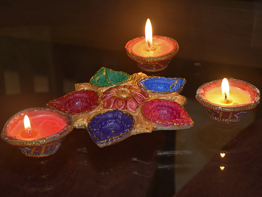 Three lit decorative oil lamps and a multi-colored decorative diya holder are placed on a reflective surface, commonly used during Diwali celebrations.
