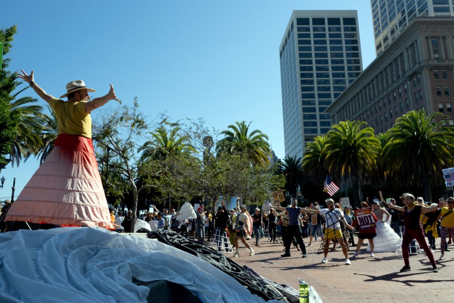 A person in a wide skirt and hat stands on a platform addressing a group of people gathered outdoors in a city plaza with palm trees and tall buildings in the background.