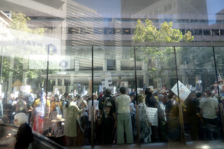 A large crowd of people holding signs gathers outside a building, seen through a glass window on a sunny day.