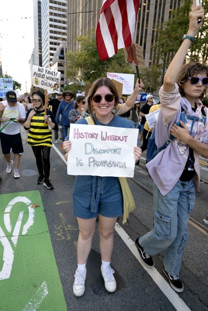 A person at a protest holds a sign reading "History without discomfort is propaganda." Other demonstrators and signs are visible in the background on a city street.