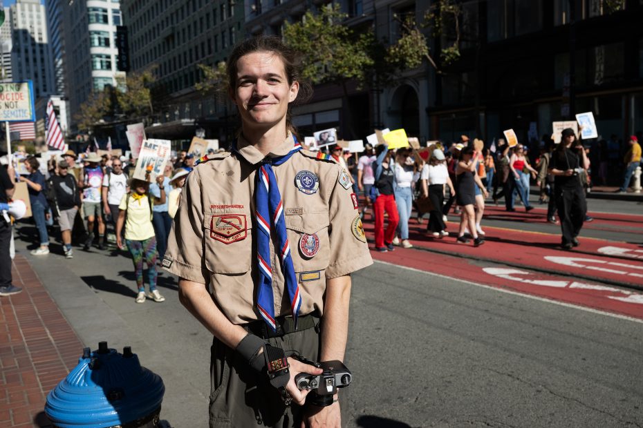 A person in a scout uniform stands on a city street, holding a camera, with a large group of protesters carrying signs in the background.