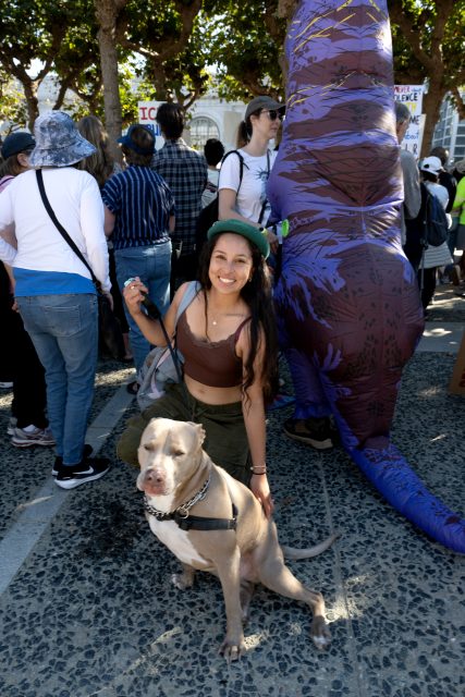 A woman kneels next to a large dog on a leash at an outdoor gathering with people and protest signs visible in the background.