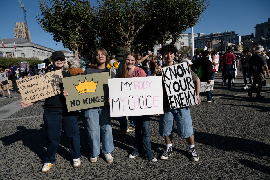 Four people stand outdoors at a protest holding signs that read: "Immigrants make America great," "No kings," "My body, my choice," and "Know your enemy.
