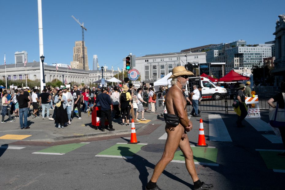 A man wearing only a cowboy hat, sunglasses, and minimal clothing walks across a crosswalk at a busy urban outdoor event with a crowd and city buildings in the background.